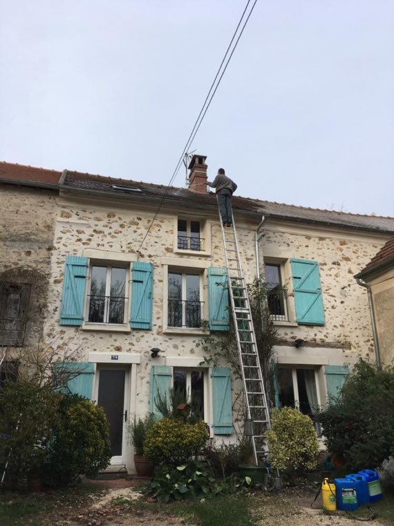Échelle de couvreur dressée le long d'une maison en pierre à volets bleus, ramoneur sur le toit